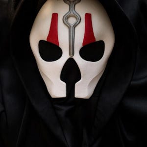 Könnte beinhalten: Eine weiße Vollgesichtsmaske mit einem silbernen vertikalen Emblem auf der Stirn, zwei roten vertikalen Markierungen über den Augenöffnungen und einer schwarzen Kapuze, die den Rand umschließt. Die Maske hat große, dunkle Augen- und Nasenausschnitte.