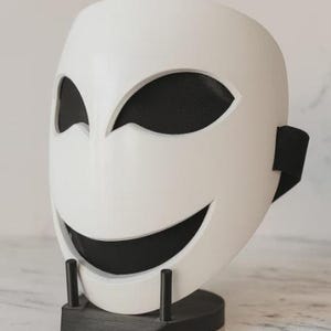 May include: A white mask with a smiling face design, featuring black mesh eye and mouth openings. The mask is displayed on a black stand. The mask has a black strap on the side.