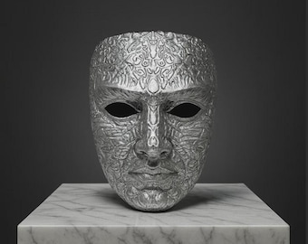 May include: A silver-colored mask with intricate, embossed patterns. The mask has a human face with empty eye sockets and is displayed on a marble-topped pedestal against a dark gray background. The mask's surface is detailed with swirling designs.