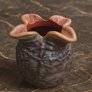 May include: A small, rounded ceramic vessel with a textured, dark purple and grey exterior surface. The rim features four distinct, petal-like lobes that curve outward, revealing a smooth, reddish-brown interior finish within the opening.