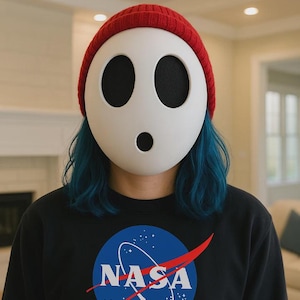 Puede incluir: Una persona con una máscara blanca con agujeros negros para los ojos y un gorro rojo. La persona lleva una sudadera negra de la NASA con un logotipo azul y blanco y un trazo rojo.