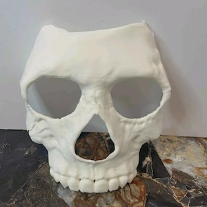 Puede incluir: Una máscara de calavera blanca con grandes cuencas oculares, una abertura nasal triangular y una mandíbula detallada. La máscara está hecha de un material sólido y está diseñada para ser usada. El tema general es Halloween o disfraz.