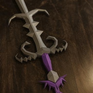 May include: A papercraft sword with a silver blade and guard, and a purple handle and pommel. The sword is lying on a dark wood surface. The design features sharp angles and geometric shapes.