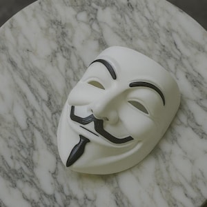 May include: A white mask with black accents, featuring a stylized smile, mustache, and eyebrows. The mask is resting on a marble-patterned surface, creating a contrast between the smooth mask and the textured background. The mask is a symbol of anonymity.