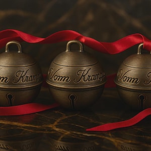 Puede incluir: Tres campanas de color bronce con la inscripción "Vom Kramp" están unidas por una cinta roja. Cada campana tiene un diseño de ojo de cerradura y un lazo. El fondo es una superficie oscura y texturizada.