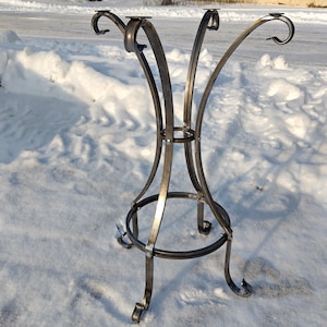 May include: A hand-forged iron table base with a dark, metallic finish. The base has curved legs and a circular support ring, designed to hold a tabletop. The top has four curved supports, each with a flat surface for attaching a tabletop.