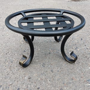 May include: Black metal plant stand with a circular top and a woven pattern. The stand has three curved legs with decorative, gold-coloured accents. The stand is designed to hold a potted plant and is suitable for indoor or outdoor use.