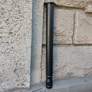 Peut inclure: Un pied de meuble noir, de forme cylindrique avec une base arrondie décorative. Le pied est en métal et est fixé à une plaque carrée en haut. Le pied est présenté contre un mur de pierre grise texturée.