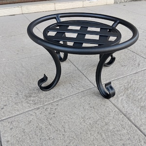 Könnte beinhalten: Ein schwarzer Pflanzenständer aus Metall mit einer kreisförmigen Oberseite und geschwungenen Beinen. Der Ständer hat ein Gitterdesign auf der Oberseite und dekorative Verzierungen an den Füßen. Für einen Blumentopf.