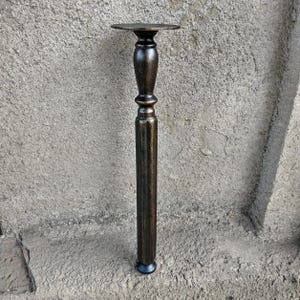 Può includere: Una gamba per mobili alta di colore marrone scuro con una finitura metallica color bronzo. La gamba presenta una piastra di montaggio circolare piatta sulla parte superiore, una sezione centrale liscia e bombata e un fusto inferiore scanalato.