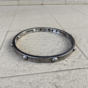 May include: A circular, silver-toned metal object with a flat band and rounded rivets. The object is set against a neutral, light-colored background. The metal has a slightly worn appearance.