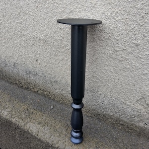 May include: A black furniture leg with a round top and decorative turned details. The leg is made of metal and is approximately 12 inches tall.