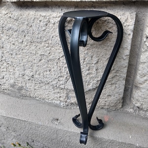 May include: A black metal plant stand with a round top and decorative, curved legs. The stand has a heart-shaped design and is suitable for displaying small plants or decorative items. The stand is approximately 24 inches tall.