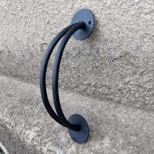May include: Black metal handrail with a double loop design. The handrail is mounted to a wall with two round plates.