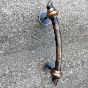 May include: A decorative door handle with a bronze and black finish. The handle has a curved design with a textured appearance and is mounted on a gray stone surface. The handle is approximately 10 inches long.
