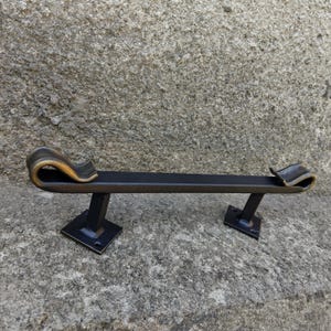 May include: A black metal door pull with decorative, curved ends. The ends have a gold-colored accent. The door pull is mounted on two square black metal bases.