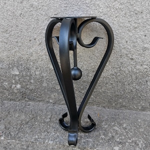 May include: Black wrought iron wall-mounted shelf with a heart-shaped design. The shelf has a small, round top and decorative scrollwork at the base. A small, spherical detail hangs from the center.