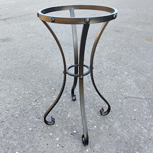 Op de afbeelding: Een donker metalen plantenstandaard met een cirkelvormige bovenkant en gebogen poten. De standaard heeft een decoratieve ring rond de poten en is ontworpen om een ​​potplant te bevatten. De standaard is gemaakt van metaal en heeft een donkere afwerking.