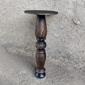 May include: A tall, slender pedestal stand featuring a flat, circular top and a decorative, carved wooden column. The central support displays a dark brown, stained finish with a spiraling, twisted texture and rounded, bulbous segments.