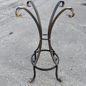 May include: A black wrought iron table base with four curved supports. The supports have decorative gold-colored accents at the top and bottom. The base is designed to hold a round tabletop.