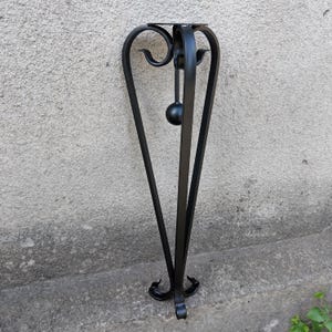 May include: A black metal plant stand with a round top and a decorative, curved design. The stand has three legs that curve outward and a small, round ball suspended in the center. The stand is approximately 30 inches tall.