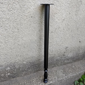 May include: A matte black metal furniture leg featuring a long, cylindrical shaft with a flat circular mounting plate at the top and a decorative, turned-style bulbous detail at the base, providing a sturdy support for tables or desks.