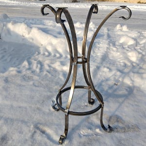 Puede incluir: Una base de mesa de hierro forjado a mano con un acabado metálico oscuro. La base presenta patas curvas y un anillo de soporte circular, diseñado para sostener una mesa. La parte superior tiene cuatro soportes curvos.