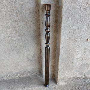 May include: A tall, slender furniture leg with a dark bronze finish and a distressed, antiqued texture. The leg features a circular mounting plate at the top, followed by a series of decorative turned, rounded, and fluted vertical segments.