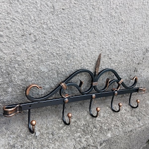 Pode incluir: Um cabide de parede de metal preto com seis ganchos. O cabide apresenta detalhes decorativos e detalhes em cobre. Os ganchos são espaçados uniformemente e têm um design arredondado. O cabide está montado numa parede cinzenta texturizada.