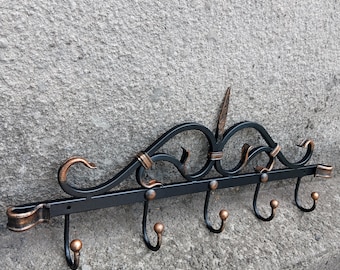 Op de afbeelding: Een zwarte metalen wandkapstok met zes haken. De kapstok heeft decoratieve krullen en koperkleurige accenten. De haken zijn gelijkmatig verdeeld en hebben een afgerond ontwerp. De kapstok is bevestigd aan een grijze, getextureerde muur.