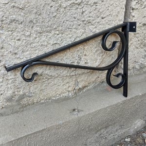 May include: A black metal wall bracket featuring a triangular frame with two decorative scrollwork elements. The bracket includes a vertical mounting plate with a visible screw hole at the top and a horizontal arm with a small upturned end hook.