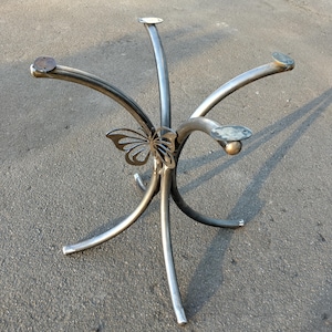 Op de afbeelding: Een metalen tafelpoot met een vlinderontwerp. De poot heeft vier poten die naar boven buigen en in het midden samenkomen. De vlinder is uit het metaal gesneden en bevindt zich in het midden van de poot.