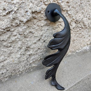 May include: Black cast iron door knocker in a leaf design. The knocker is mounted on a textured, light grey wall. The leaf-shaped knocker has detailed veining and a curved handle.