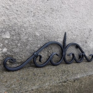 Op de afbeelding: Zwart smeedijzeren decoratieve wandkunst met een krulontwerp. Het metalen kunstwerk heeft gebogen lijnen, krullen en een centraal verticaal element. Ontworpen voor wandmontage, voegt het een vleugje elegantie toe.
