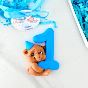 May include: A brown teddy bear figurine wearing a blue party hat, holding a large blue number one. A tag with the name "Oliver" and the date 06/06/2025 is attached with a blue ribbon. The background includes blue paper shreds.