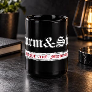Könnte beinhalten: Ein schwarzer zylindrischer Becher mit weißem Text im gotischen Stil, der oben "Sturm & Drang" lautet. Darunter befindet sich ein weißer horizontaler Pinselstrich-Streifen, der den roten Text "Recht auf Meinung" in gotischer Schrift enthält.