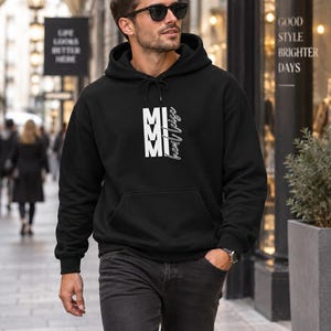 Könnte beinhalten: Ein schwarzer Kapuzenpullover mit einer weißen Grafik auf der Brust, bestehend aus den vertikal gestapelten Buchstaben MMI und dem kursiven Schriftzug 'hem leise' rechts daneben. Das Kleidungsstück hat eine Kängurutasche und lange Ärmel.