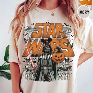 May include: An ivory t-shirt featuring a graphic of Darth Vader wearing Mickey Mouse ears and holding a jack-o'-lantern. The design includes the text STAR WARS, stormtroopers with mouse ears, spaceships, and bats in orange and black ink.