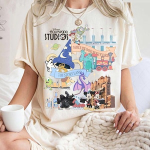Può includere: T-shirt color crema con una grafica colorata di Disney's Hollywood Studios. Il design include personaggi di Toy Story, Monsters & Co. e Mickey Mouse, con testi come "Hollywood Boulevard" e "Toy Story Land."