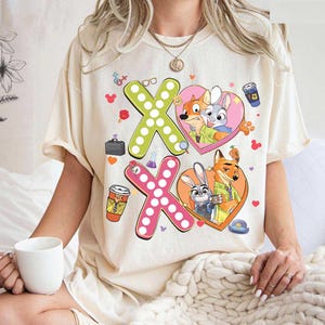 May include: Cream-colored t-shirt featuring a graphic design with the characters from Zootopia. The design includes two "XO" shapes, each with a heart-shaped image of the characters. Additional cartoon elements are scattered around the design.
