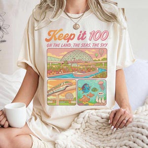 Puede incluir: Camiseta color crema con un gráfico colorido. El diseño incluye el texto "Keep it 100" e ilustraciones de un paseo en barco, un tren y una escena submarina. La camiseta tiene mangas cortas y cuello redondo.