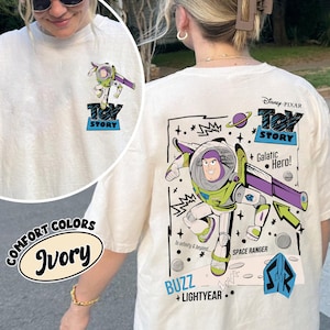 K&ouml;nnte beinhalten: Elfenbeinfarbenes Langarm-T-Shirt mit Buzz Lightyear aus Toy Story. Die Vorderseite hat eine kleine Grafik, w&auml;hrend die R&uuml;ckseite ein gr&ouml;&szlig;eres Design mit dem Text "Toy Story", "Galactic Hero" und "Buzz + Lightyear" zeigt. Das Shirt ist Teil der "Comfort Colors"-Kollektion.