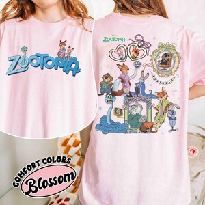 Puede incluir: Camiseta rosa claro con la palabra "Zootopia" en azul y personajes de dibujos animados. La parte trasera de la camiseta tiene gr&aacute;ficos adicionales de personajes de Zootopia. La camiseta est&aacute; etiquetada como "Comfort Colors Blossom".