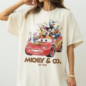 May include: Cream-colored t-shirt featuring a red cartoon car with Disney characters, including Mickey Mouse, Donald Duck, and Goofy. The text "Mickey & Co. EST 1928" is printed below the car. The shirt has short sleeves and a relaxed fit.