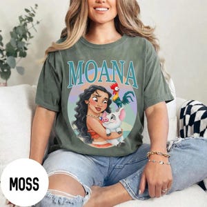 May include: Moss green t-shirt featuring the word "MOANA" in teal lettering. The shirt displays a colorful graphic of Moana, a pig, and a rooster. The shirt is a relaxed fit and has short sleeves.