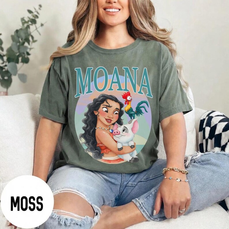 May include: Moss green t-shirt featuring the word "MOANA" in teal lettering. The shirt displays a colorful graphic of Moana, a pig, and a rooster. The shirt is a relaxed fit and has short sleeves.
