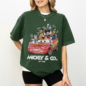 May include: Dark green t-shirt featuring a red cartoon car with Mickey Mouse and friends. The car has the number 95 on it and the text "Mickey & Co. Est 1928". The shirt is a crew neck.