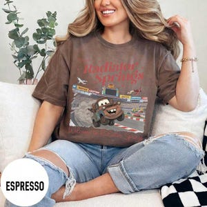 May include: Espresso-colored t-shirt featuring a graphic of Mater from the movie Cars racing on a track. The text "Radiator Springs" and "Welcome to Radiator Springs" are printed in red. The shirt is a short-sleeved, casual style.