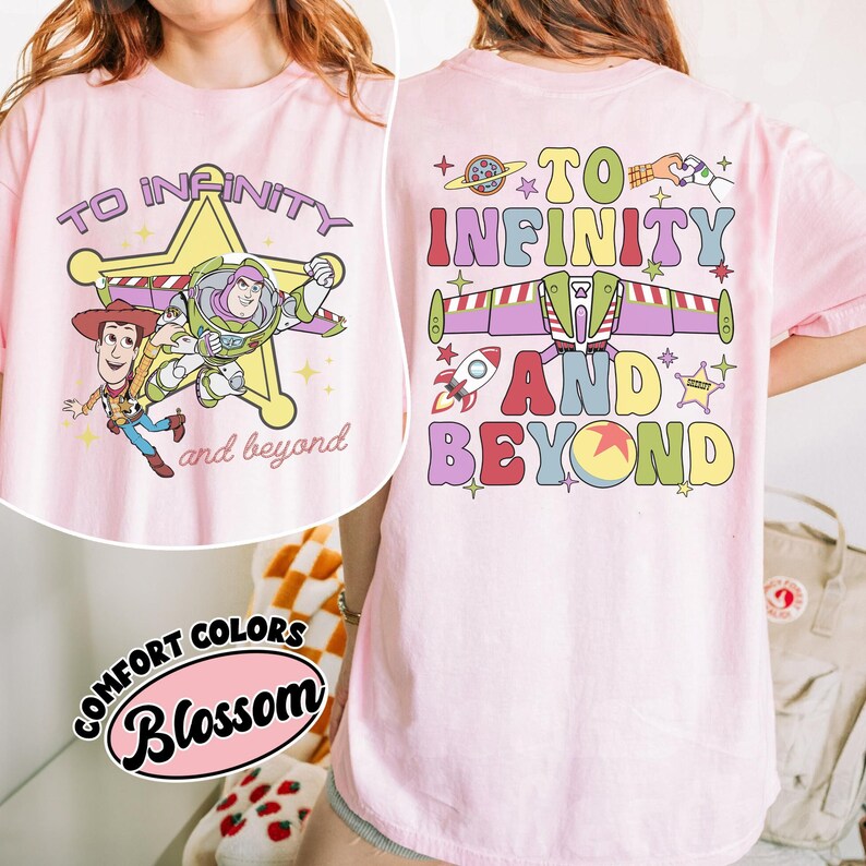 May include: Pink t-shirt featuring a graphic of Woody and Buzz Lightyear with the text "To Infinity and Beyond." The back of the shirt has a matching design with a rocket ship and the same phrase. The shirt is a Comfort Colors Blossom.