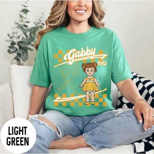 May include: Light green t-shirt with a graphic of a doll and the name "Gabby 1995" in a retro style. The shirt has a crew neck and short sleeves. The text "Light Green" is at the bottom of the image.